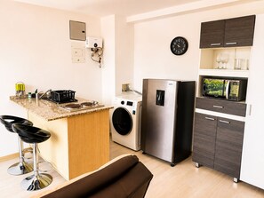 Cocina compacta con lavadora integrada, refrigerador, microondas y un reloj en la pared.