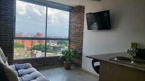 A modern living room with a large window, a flat-screen TV, a sofa, a small kitchen area, and a potted plant. - apartament with the best views  (Medellín)