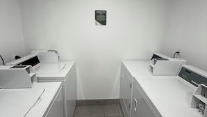 A laundry room with two washing machines and a drying machine, a sign indicating the room's purpose, and a ceiling-mounted light fixture.
