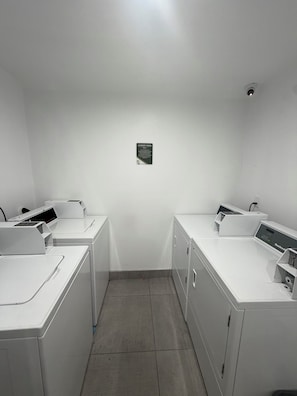 A laundry room with two washing machines and a drying machine, a sign indicating the room's purpose, and a ceiling-mounted light fixture.