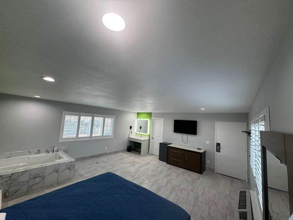 A spacious room with a large bed, a built-in bathtub, a vanity with a mirror, a television, and a window with blinds.