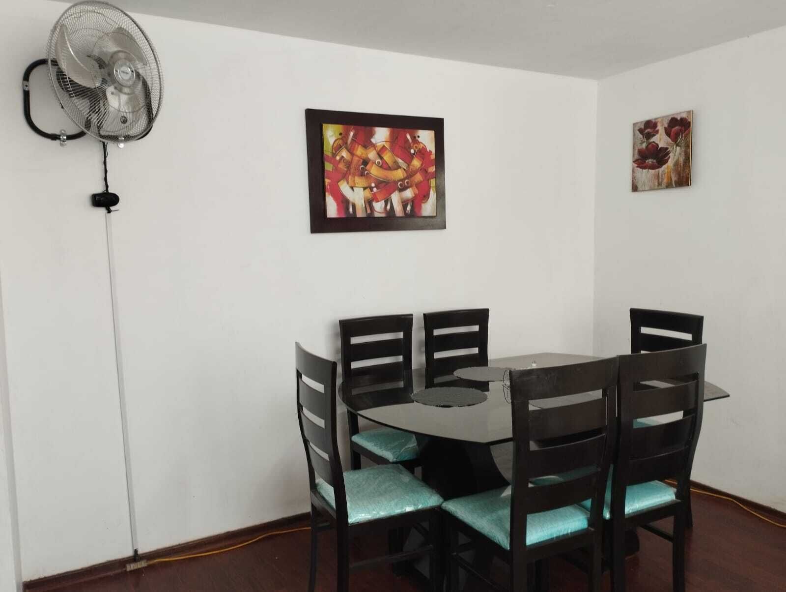 Un comedor con una mesa y sillas, un ventilador de pie y dos obras de arte enmarcadas en la pared.