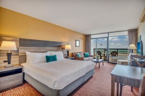 A hotel room with a large bed, two bedside tables with lamps, a sofa, a small table, and a balcony with a view of the sky.