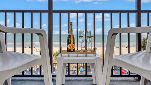 A balcony with a view of the beach, featuring a bottle of champagne and two glasses on a small table. - Oceanfront Sea and Sky Haven! (Myrtle Beach)