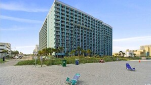 A large hotel with multiple floors and balconies, situated on a beach with a few beach chairs and a trash bin. - Oceanfront Sea and Sky Haven! (Myrtle Beach)