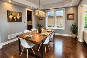 A dining room with a wooden table set for six, white chairs, a chandelier, and a window with a view of a brick wall. - 8BR Estate by Canterra Stays · Sleeps 20 · Games (Brampton)