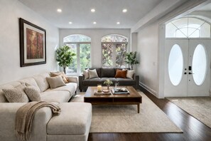Ein geräumiges Wohnzimmer mit einem beigefarbenen Sofa, einem dunkelgrauen Sofa und einem Holztisch. Zwei große Fenster mit Rundbögen lassen natürliches Licht herein.
