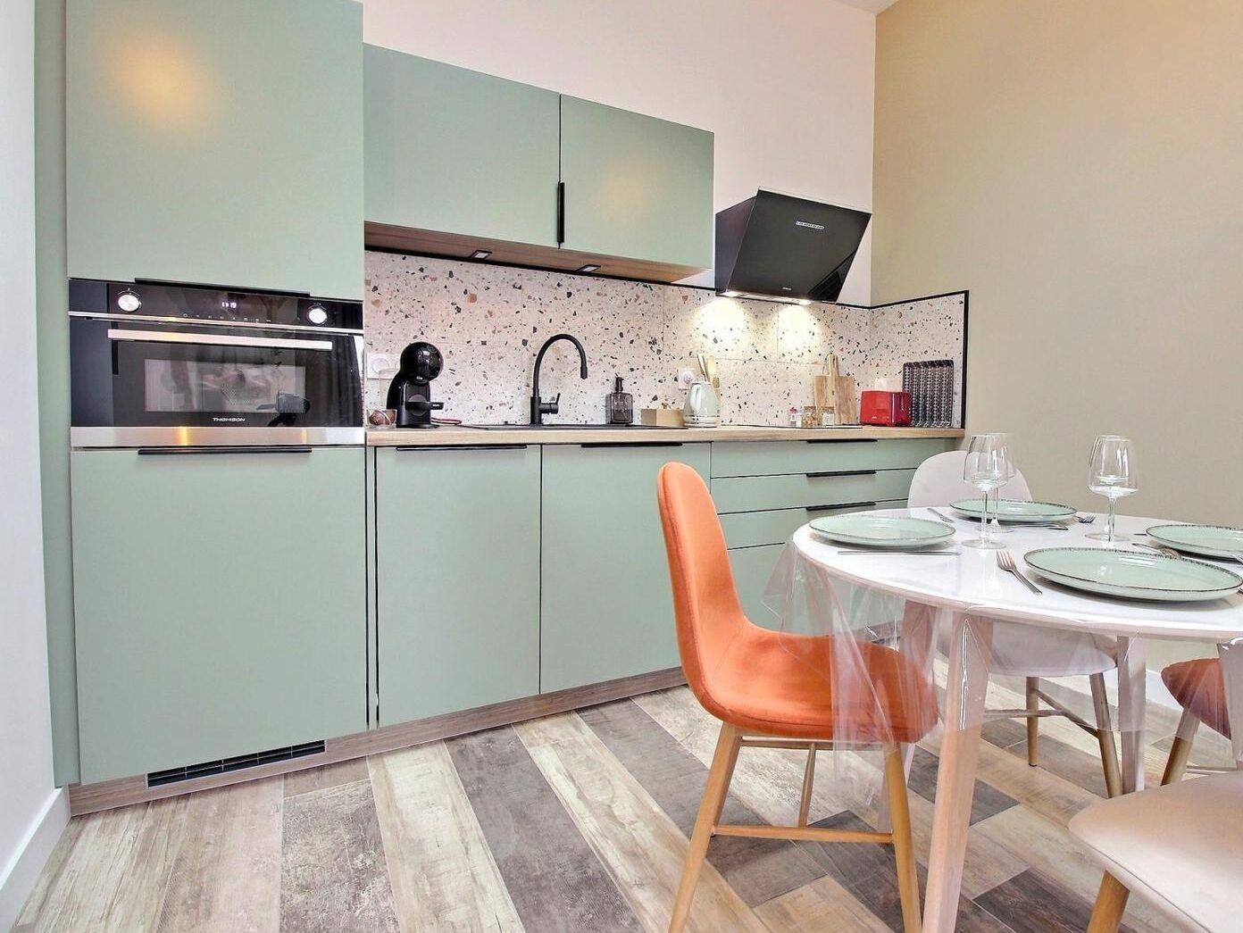 A compact kitchen with a microwave, kettle, and sink. There is a dining table with chairs and a glass of wine.
