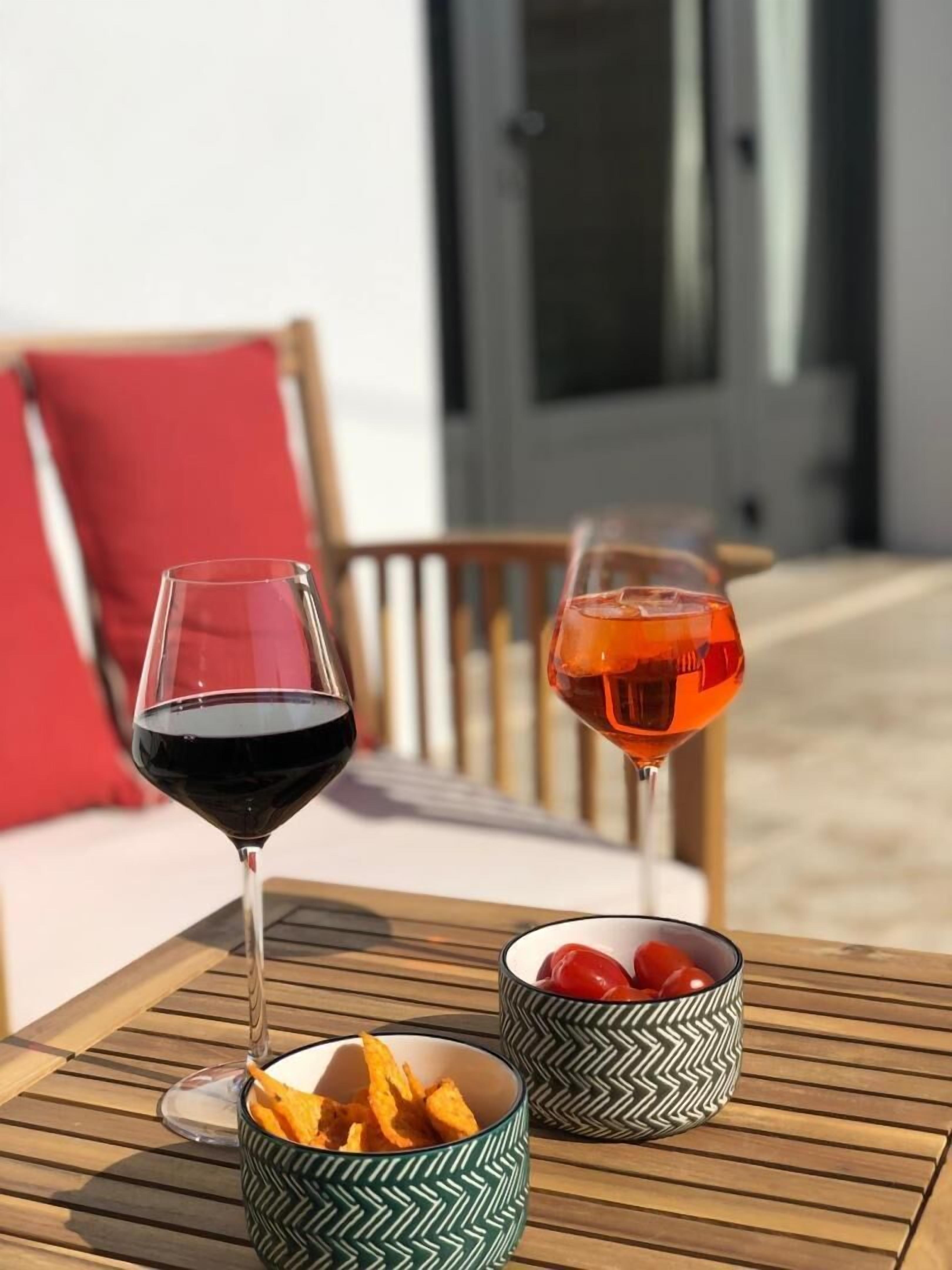Two wine glasses on a wooden table, one with red wine and the other with rosé, and two patterned bowls with snacks and tomatoes.
