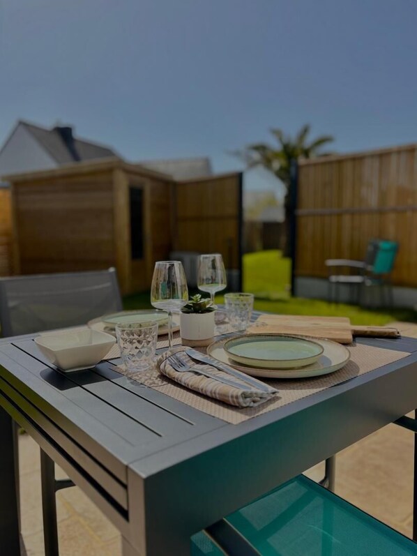 An outdoor dining table set with plates, glasses, and cutlery, surrounded by a wooden fence and a chair.