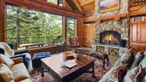 L’intérieur chaleureux d’un chalet avec une cheminée en pierre, des meubles en bois et une grande fenêtre donnant sur une forêt.