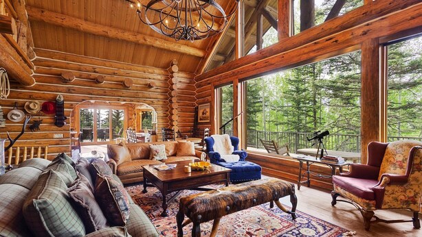 L’intérieur d’une cabane en rondins, avec une grande fenêtre donnant sur une forêt, un coin salon confortable avec un canapé et des fauteuils, et un plafond en bois avec des poutres apparentes.