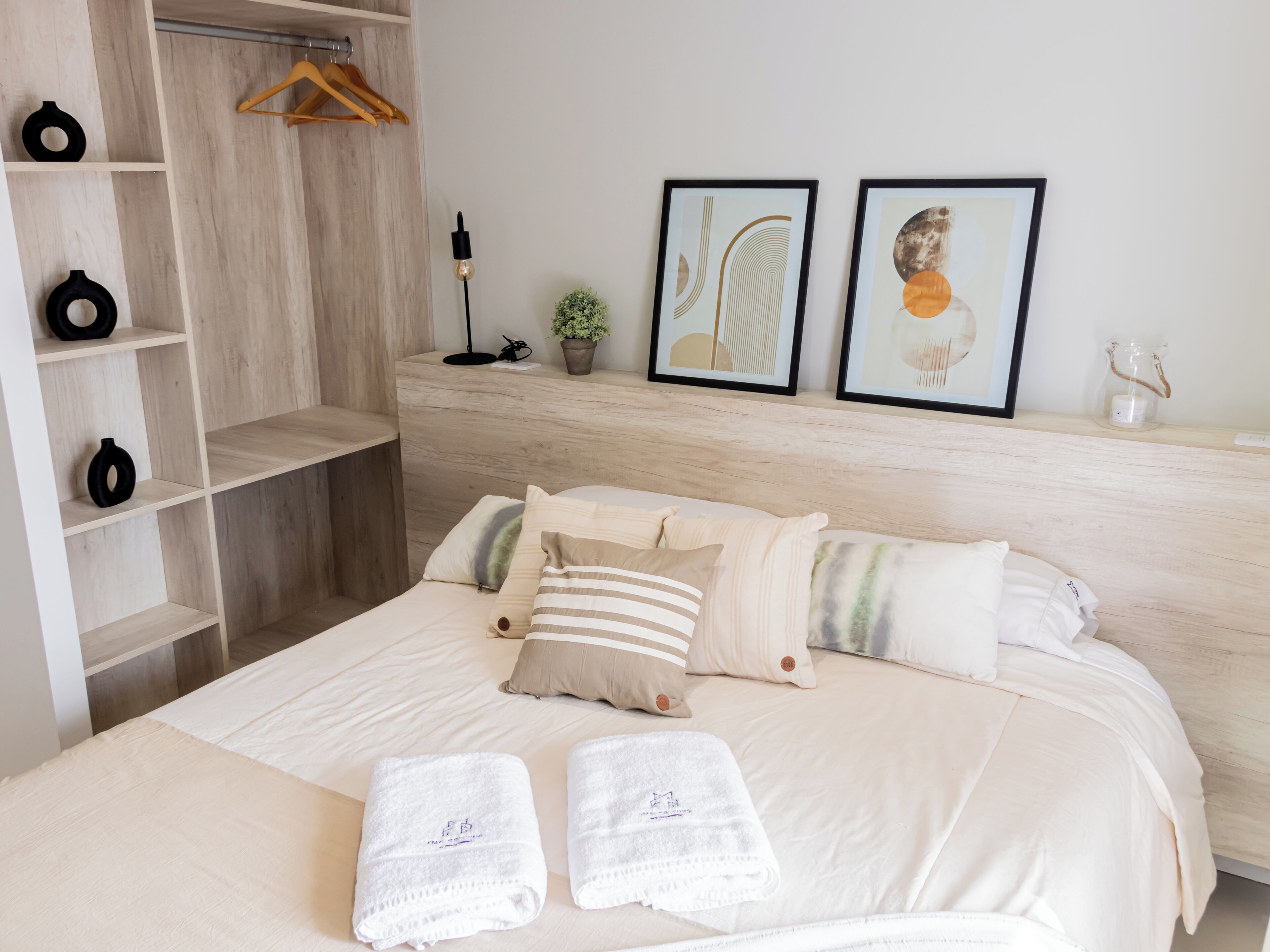 Un dormitorio ordenado con cama, almohadas, una estantería con objetos decorativos y dos cuadros en la pared.