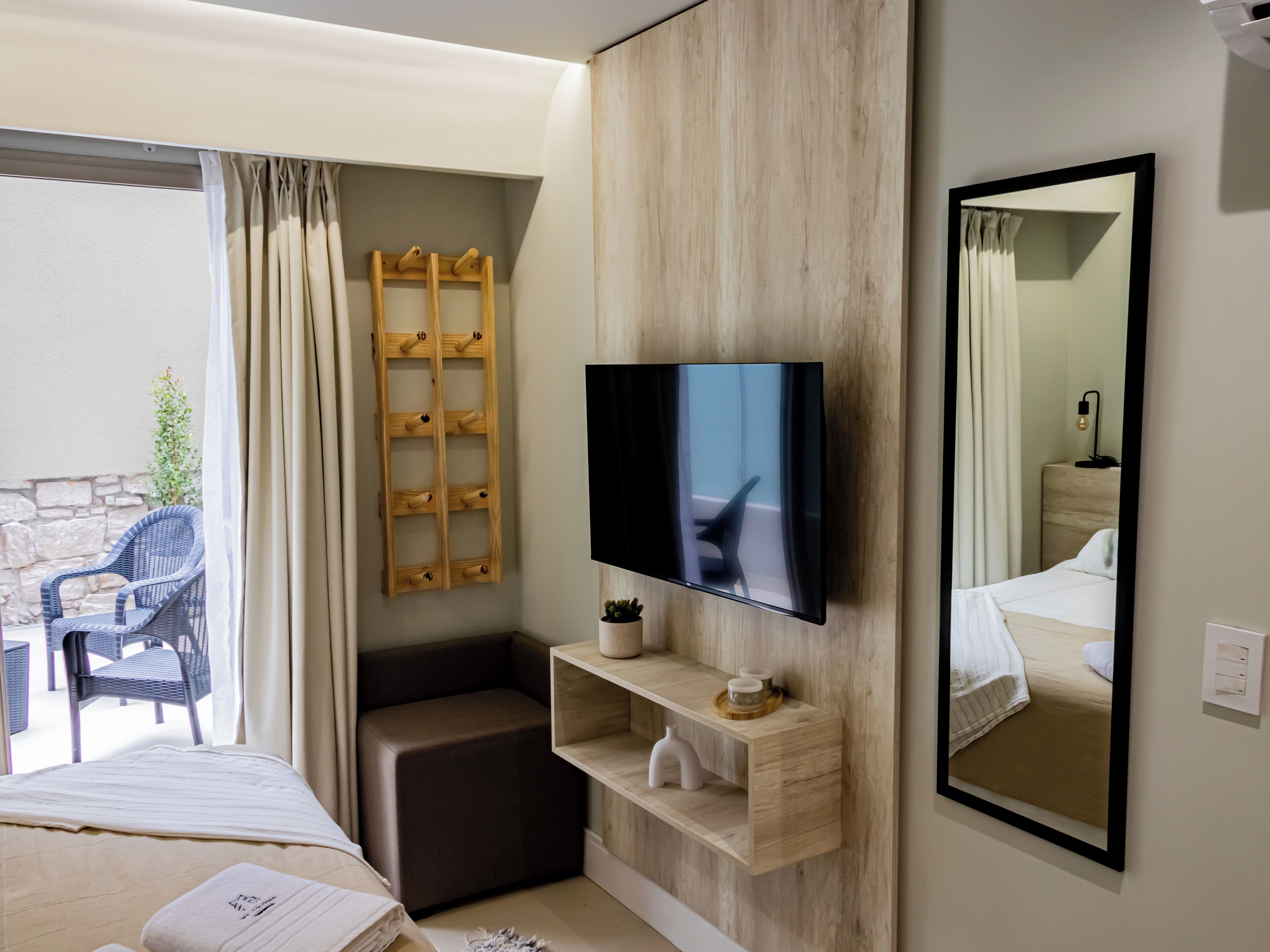 Una habitación de hotel moderna con una cama, un televisor montado en la pared, un estante de madera con artículos de aseo, un espejo y vistas al área de estar exterior.