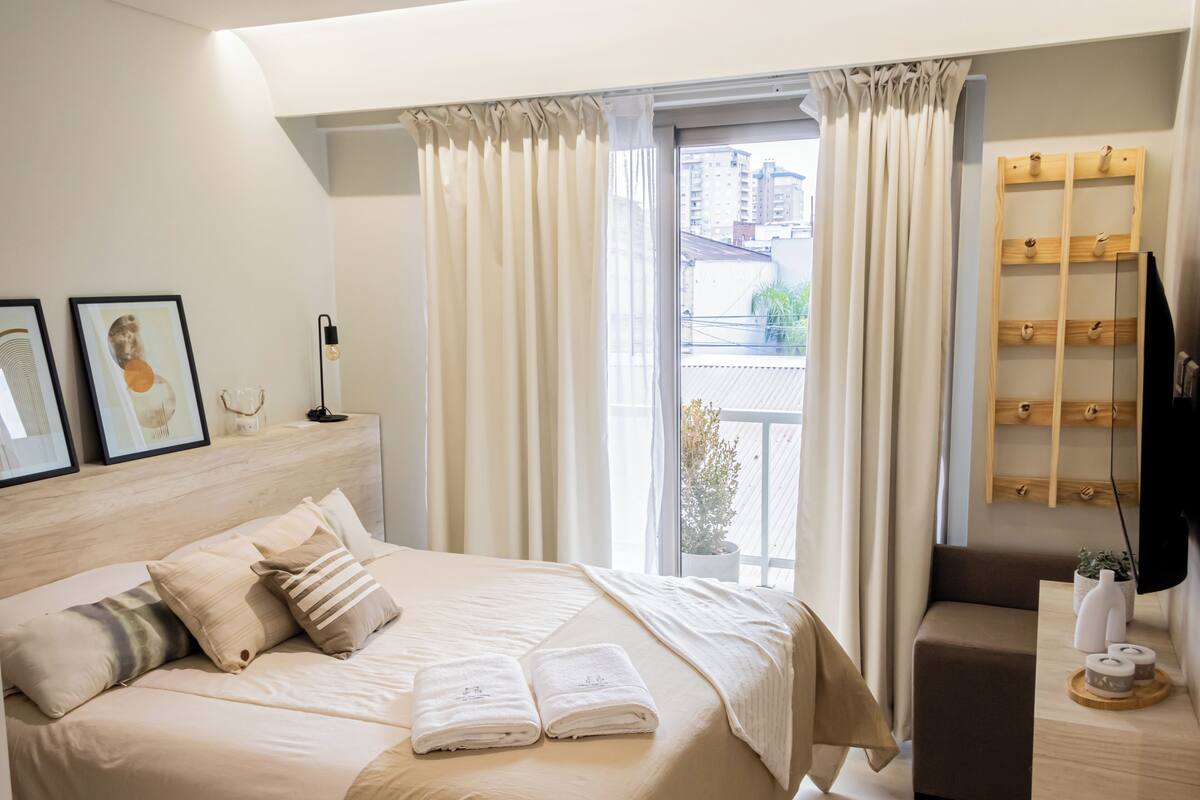 A modern bedroom with a large bed, a bedside table, a TV, and a view of the city.