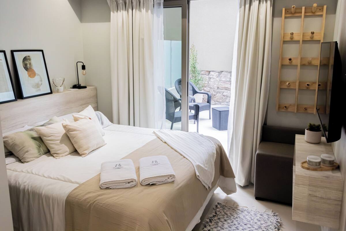 A modern hotel room with a large bed, a desk, a TV, and a balcony with outdoor seating.