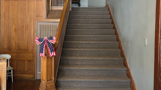 Un escalier avec des rampes en bois et des marches recouvertes de moquette, menant à un palier orné d’un cadre et d’une applique murale.