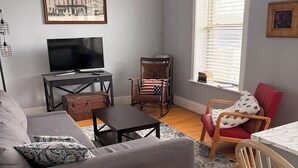 Un salon avec un canapé, un téléviseur, une table en bois et une fenêtre avec des stores.