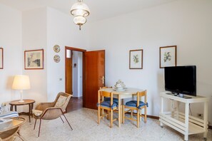 A room with a dining table, chairs, a television on a stand, a lamp, and framed pictures on the walls.
