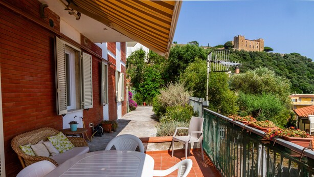 Un balcón con mesa y sillas, vista a un castillo en una colina, y un jardín con plantas en macetas.