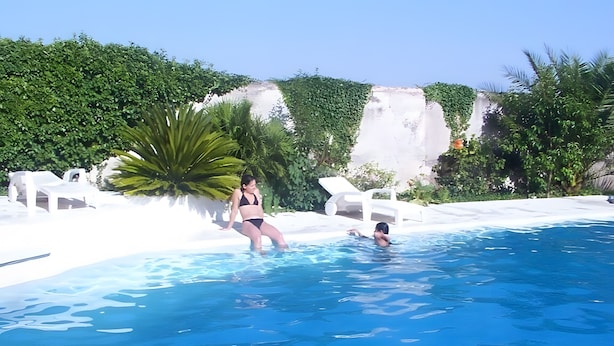En swimmingpool med hvide liggestole og grøn vegetation i baggrunden.