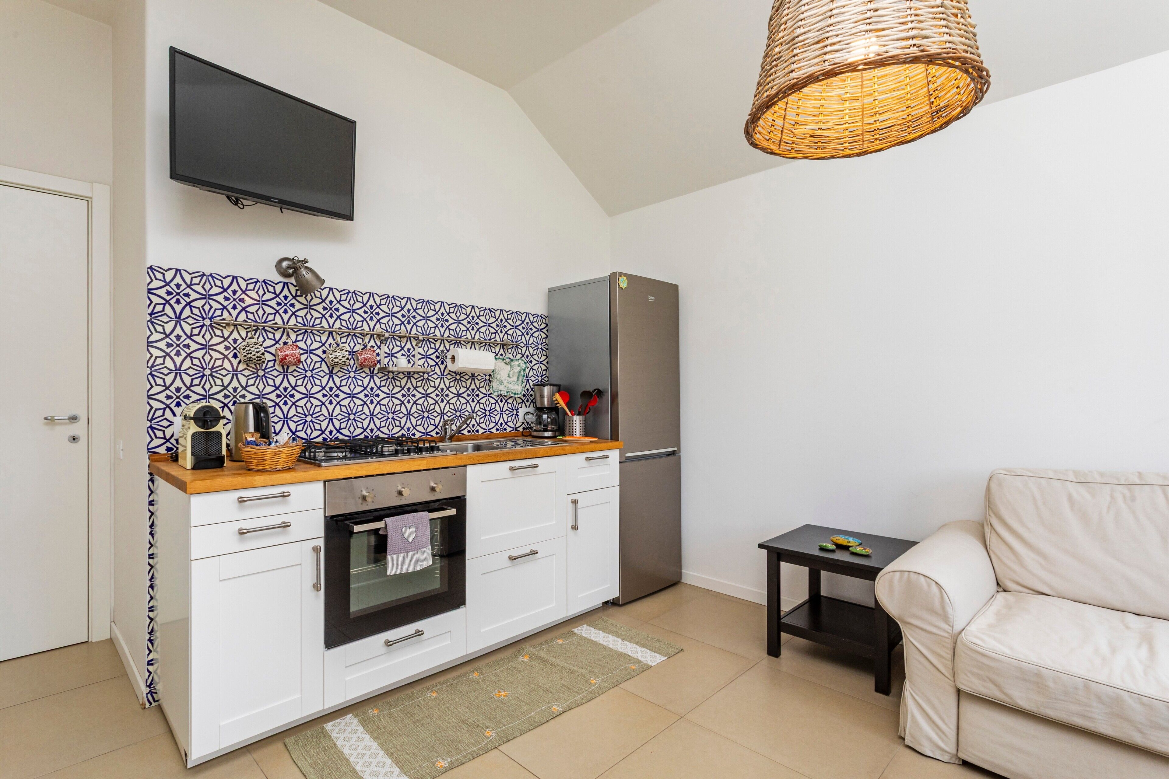 A compact kitchen with white cabinets, a stainless steel refrigerator, a flat-screen TV, and a wicker ceiling light fixture.