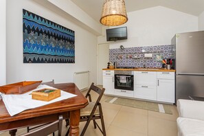 A kitchen with white cabinets, a stainless steel refrigerator, a wooden dining table with a box, and a hanging wicker light fixture.