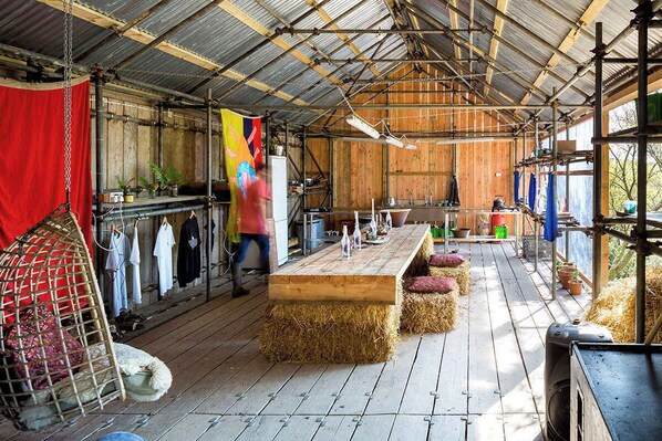 A rustic barn-style interior with a long wooden table, hay bales, and hanging clothes.