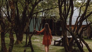 A woman walking through a wooded area with trees and a wooden structure in the background.