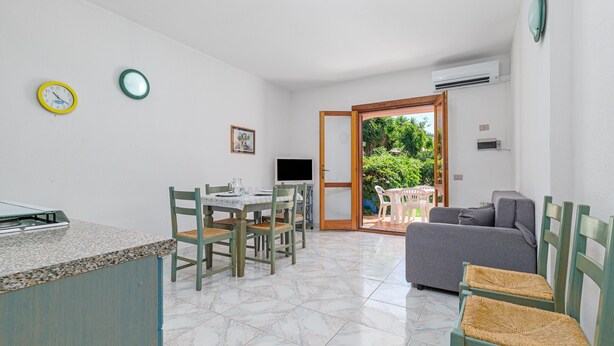 A kitchen with a dining table, chairs, a TV, and a sofa in a bright room with a tiled floor and a door leading outside. - Apartment 'Deledda' with Private Terrace, Private Garden and Air Conditioning (San Teodoro)