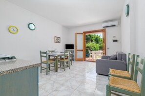 Una cucina con tavolo e sedie, TV e divano in un ambiente luminoso con pavimento piastrellato e una porta che dà all'esterno.