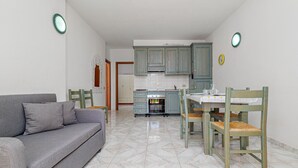 A compact living space with a sofa, dining table, chairs, kitchen area, and a clock on the wall. - Apartment 'Deledda' with Private Terrace, Private Garden and Air Conditioning (San Teodoro)
