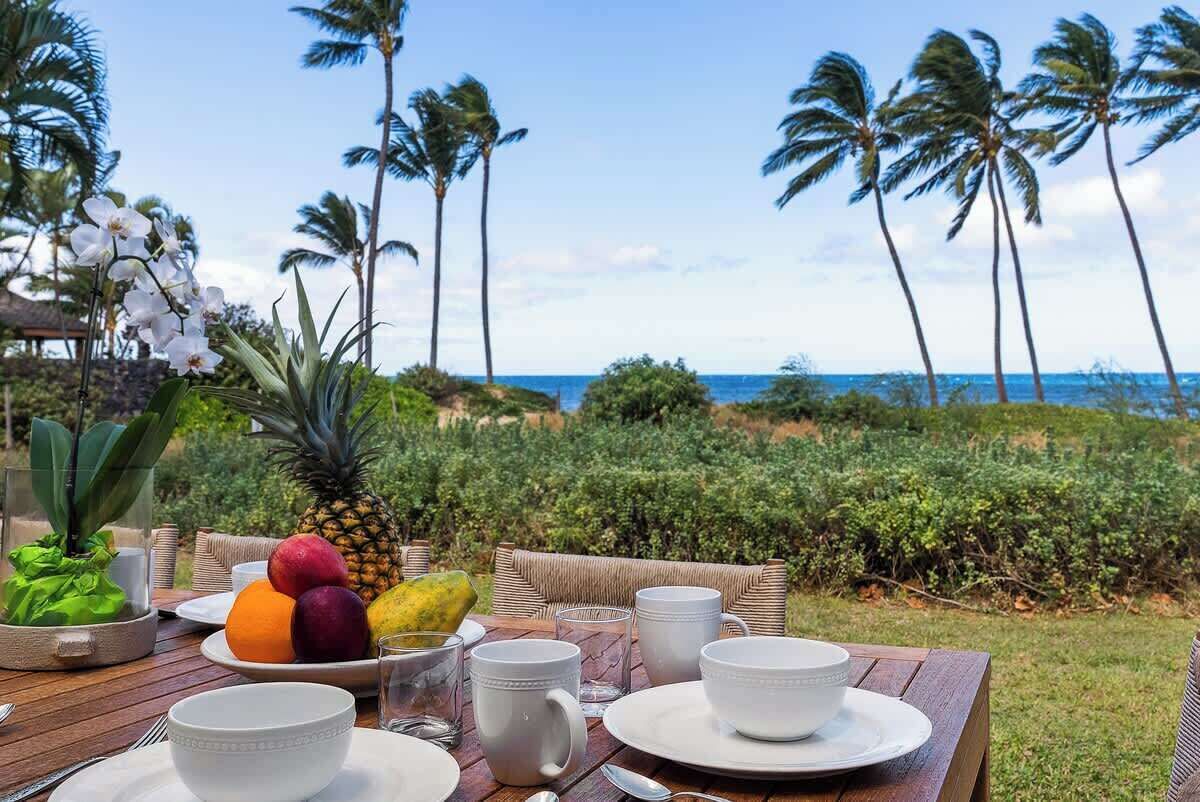 Une table dressée pour le petit-déjeuner, avec des fruits, des tasses blanches et un verre de jus, donnant sur une plage bordée de palmiers.