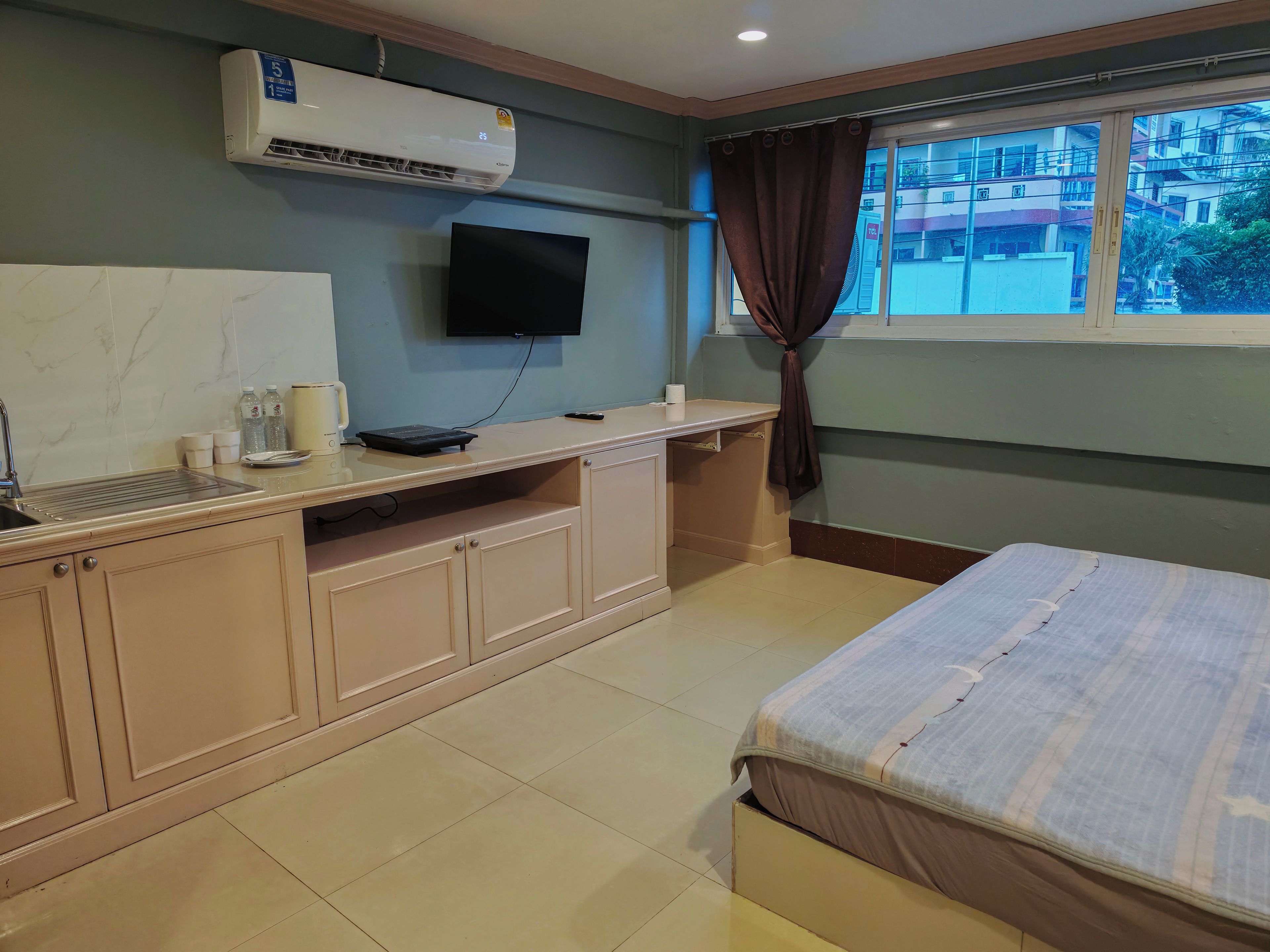A hotel room with a bed, a desk with a flat-screen TV, an air conditioner, and a window with curtains.