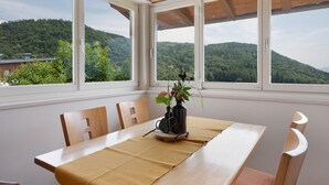 A dining area with a table, chairs, and a view of a forested hillside through large windows. - Holiday Apartment 'Schwalbennest' with Mountain View, Wi-Fi and Air Conditioning (Appiano sulla Strada del Vino)