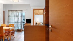 A wooden door leading to a room with a dining area, a television, and a visible kitchen area.