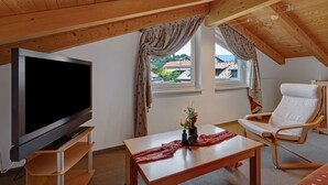 Una habitación con una televisión sobre un mueble de madera, una mesa con un florero, una silla y una ventana con cortinas.