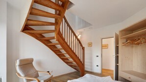 Una habitación con una escalera espiral de madera, una silla de madera, una cama con una colcha blanca y un armario de madera con perchas.