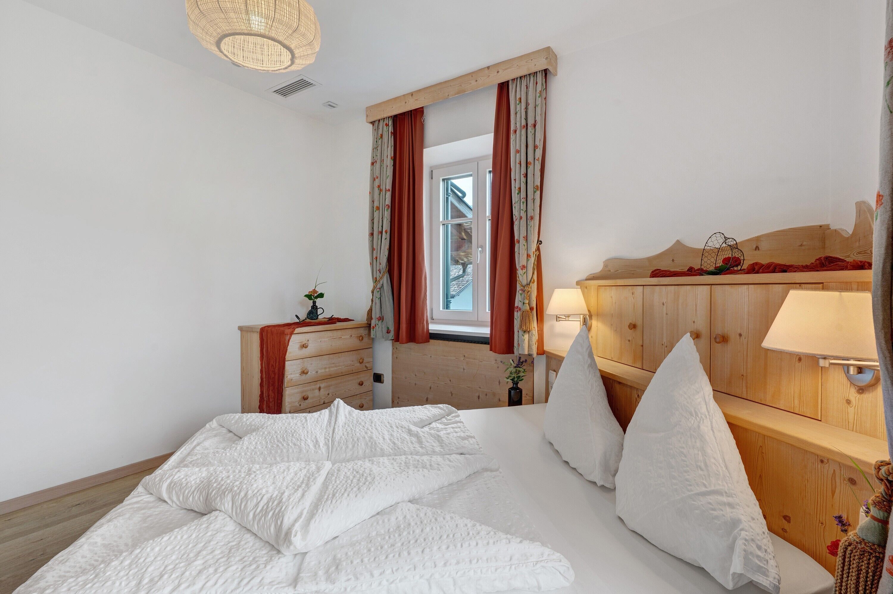 A bedroom with a wooden headboard, a bed with white bedding, a nightstand with a lamp, and a window with curtains.