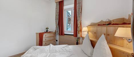 A bedroom with a wooden headboard, a bed with white bedding, a nightstand with a lamp, and a window with curtains.