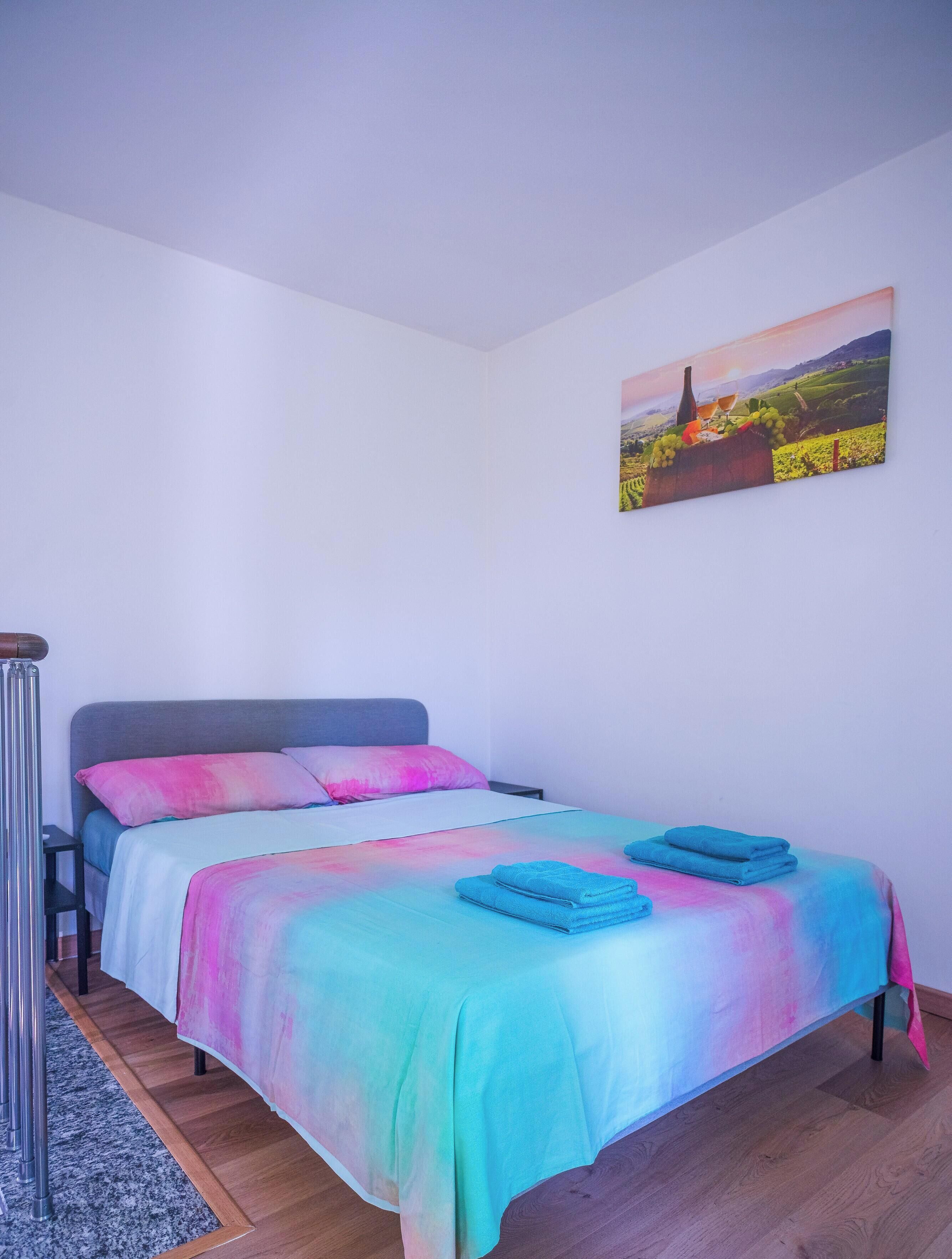 A bedroom with a bed, pillows, a bedside table, a nightstand, a wall-mounted painting, and a wooden floor.
