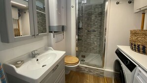 A compact bathroom with a white sink, wooden toilet, glass shower enclosure, and a washing machine.