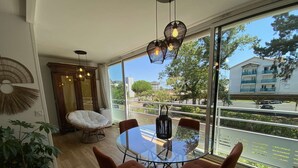 A dining area with a glass table, four chairs, a hanging light fixture, and a view of the street and buildings outside.