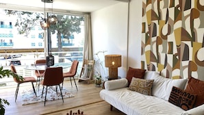 A modern living room with a sofa, a dining table, chairs, and a large window with a view of buildings and trees.