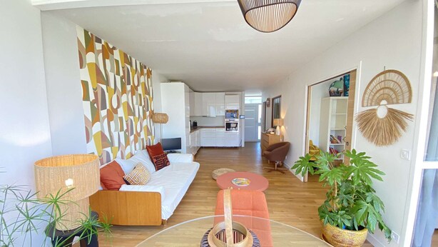 A modern living room with a sofa, coffee table, and plants. There is a kitchen in the background with white cabinets and a dining area.