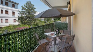 A balcony with a table and chairs, a large umbrella, and a view of buildings and trees.