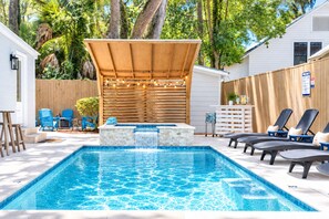 A swimming pool with a waterfall feature, surrounded by lounge chairs and a wooden deck. - Porch Light Cottage - Historic District Walk to the Beach, Private Pool, Spa, Fire Pit & Guest House (Saint Simons Island)