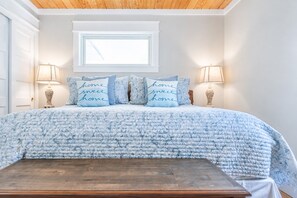 A bedroom with a bed, two bedside lamps, a window, and a wooden bench. - Porch Light Cottage - Historic District Walk to the Beach, Private Pool, Spa, Fire Pit & Guest House (Saint Simons Island)