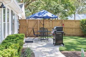 A patio with a grill, outdoor table, and chairs, surrounded by a wooden fence and greenery.