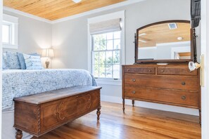 A bedroom with a wooden dresser, a bed with patterned bedding, and a window with a view of greenery. - Porch Light Cottage - Historic District Walk to the Beach, Private Pool, Spa, Fire Pit & Guest House (Saint Simons Island)